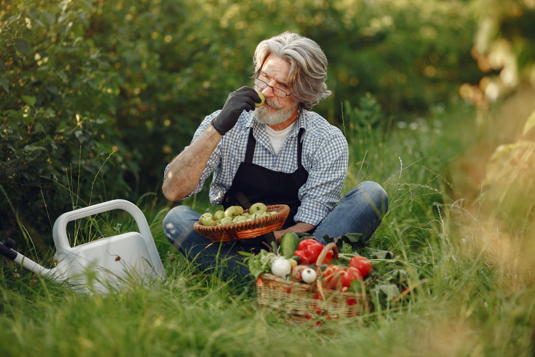 A smiling gardener sitting in long grass with a basket of tomatoes and a watering can